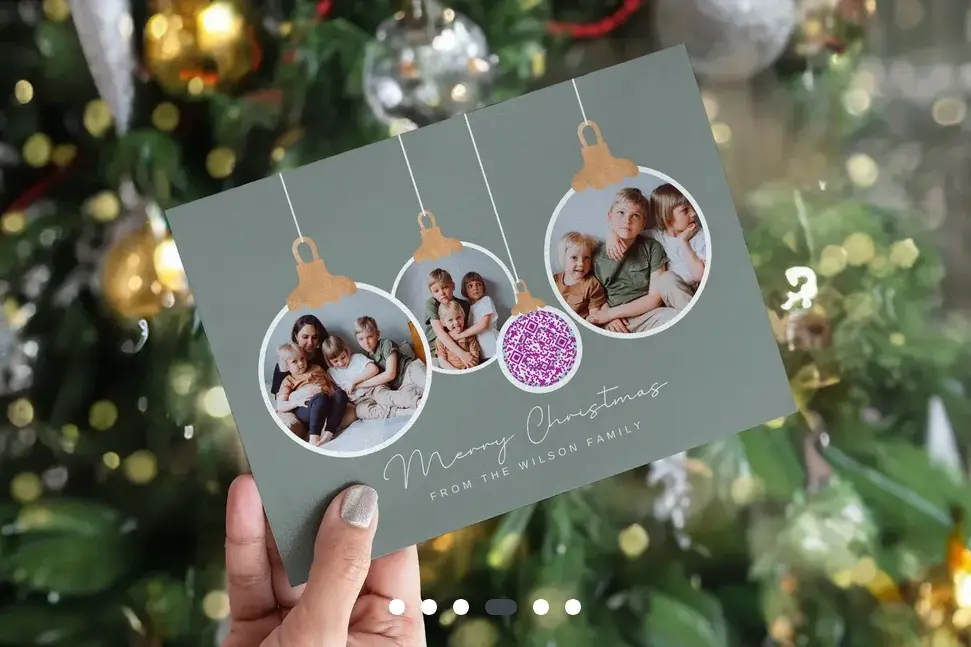 A hand with silver nail varnish holding up a family Christmas card with the words 'Merry Christmas from the Wilson Family. There are photos placed in a bauble design.