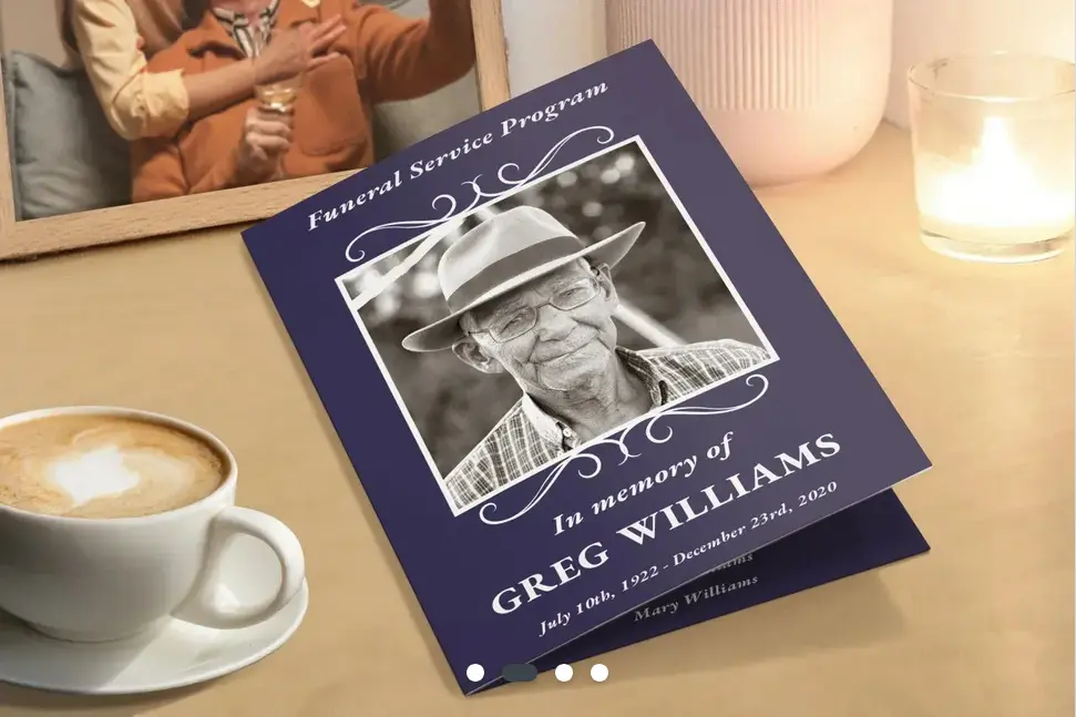 A navy blue memorial card with the photo of an elderly man in a hat. Set on a table with a coffee, a picture frame and a candle.