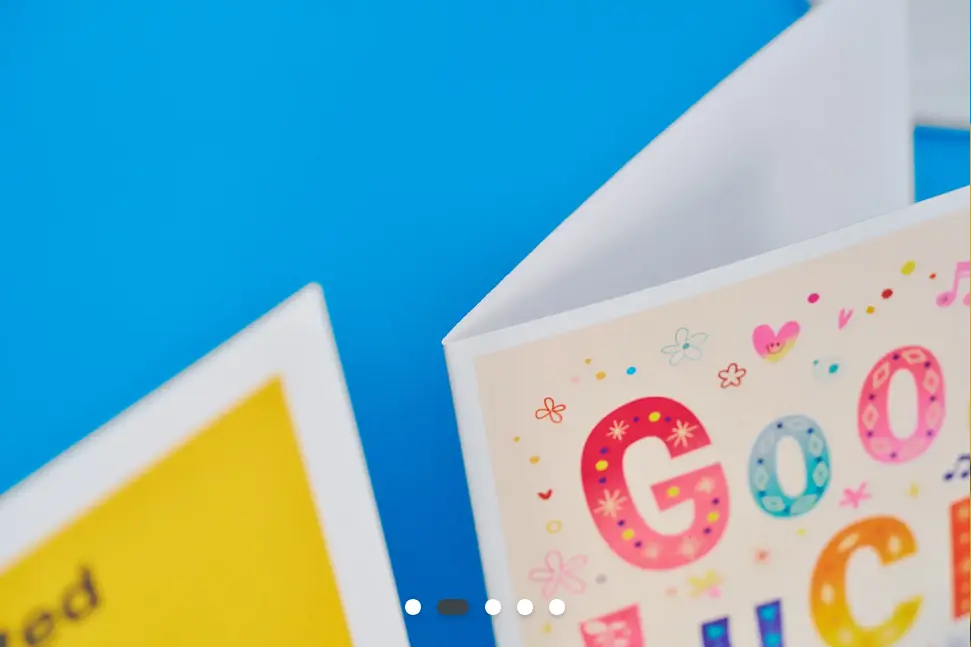 A close-up of the corner of a Good Luck Printed Card with cute flower and heart designs. There are other greetings cards in the background.